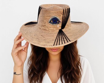 Puede incluir: Un sombrero de paja color canela con ala ancha y copa estructurada. El sombrero está decorado con un motivo central de ojo hecho de cuentas en azul, blanco y negro, acentuado por una pequeña lágrima de cristal y detalles de hilo negro.