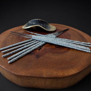 Puede incluir: Una colección de varitas de incienso de color gris claro y textura rugosa dispuestas en forma de cruz, junto a un soporte de cerámica oscuro y curvado con textura de hoja y un borde en tono dorado metálico.