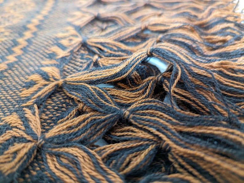 May include: A woven textile featuring a repeating geometric pattern in black and copper brown. The fabric edge displays a decorative knotted fringe detail where the black and copper brown threads are gathered and tied into distinct clusters.