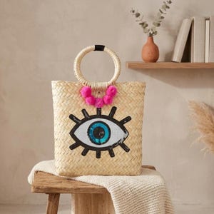 May include: A woven straw tote bag featuring a circular handle and a large embroidered eye motif on the front. The eye design includes black, white, and blue sequins, accented by a row of bright pink pom-poms attached below the handle base.