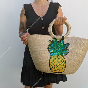 Può includere: Una grande borsa tote di paglia con un manico rotondo. La borsa ha un grande design di ananas con paillettes sul davanti. L'ananas è giallo, verde e blu.
