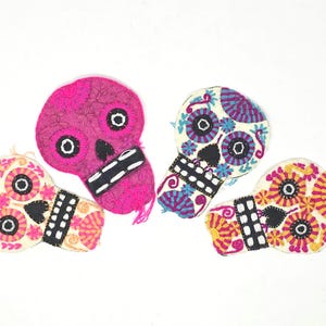 Könnte beinhalten: Vier gestickte Zucker Schädel-Ornamente aus Filz in Pink, Weiß, Blau und Gelb. Die Schädel haben schwarze Augen und Münder und sind mit Blumenmustern verziert.