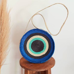 May include: A round, blue and white striped straw bag with a natural woven strap. The bag has a circular design with concentric circles of blue, white, black, and green.