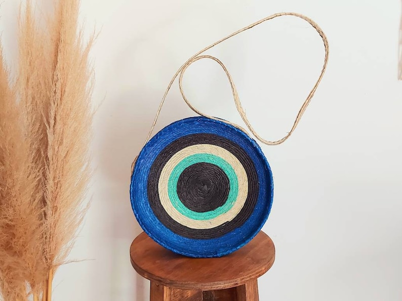 May include: A round, blue and white striped straw bag with a natural woven strap. The bag has a circular design with concentric circles of blue, white, black, and green.