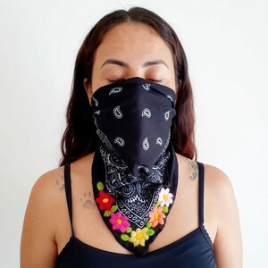 May include: A black bandana featuring a traditional white paisley pattern, worn over the nose and mouth. The lower edge of the fabric is decorated with vibrant, hand-stitched embroidery of small flowers in red, pink, orange, yellow, and green.