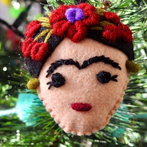 May include: A felt ornament shaped like a face with beige skin, thick black embroidered eyebrows, dark eyelashes, and red lips. The head features a black hair band adorned with red embroidered flowers and a small purple center detail.