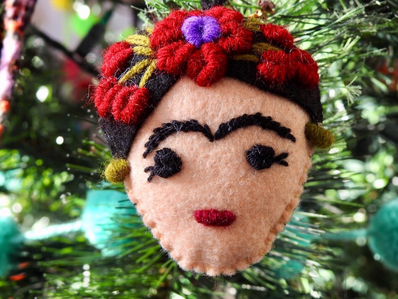 May include: A felt ornament shaped like a face with beige skin, thick black embroidered eyebrows, dark eyelashes, and red lips. The head features a black hair band adorned with red embroidered flowers and a small purple center detail.
