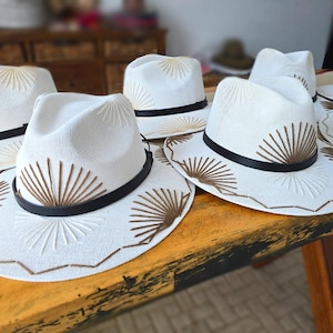 May include: A collection of white fedora hats featuring a black band and brown embroidered sunburst patterns on the crown and brim. Each hat has a structured shape with a pinched crown and a wide brim, showcasing detailed stitching throughout.