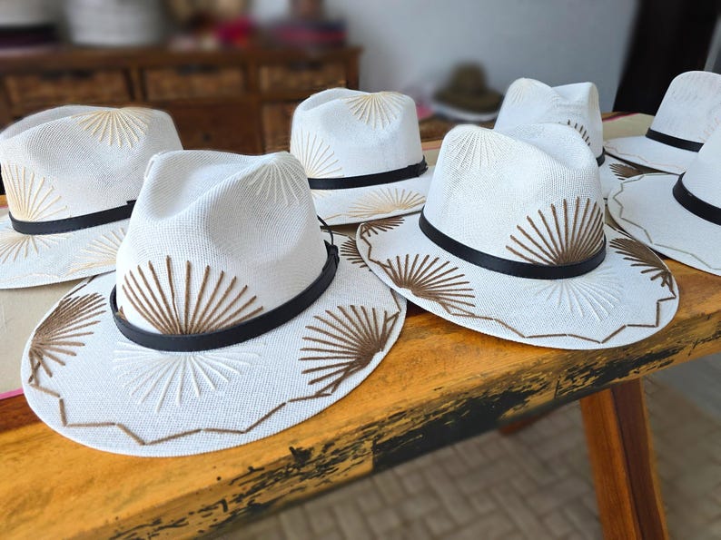 May include: A collection of white fedora hats featuring a black band and brown embroidered sunburst patterns on the crown and brim. Each hat has a structured shape with a pinched crown and a wide brim, showcasing detailed stitching throughout.