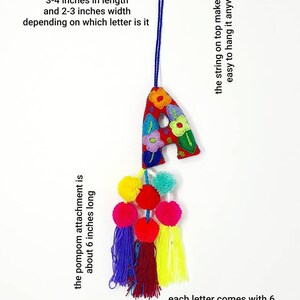 May include: A colourful felt letter A with embroidered flowers, a blue string for hanging, and a tassel of six multi-coloured pompoms. The letter measures 3-4 inches in length and 2-3 inches in width. The pompom attachment is about 6 inches long.