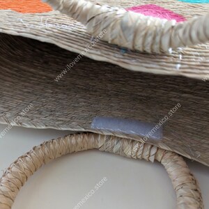 May include: A woven straw tote bag featuring two round, rigid handles and a small rectangular velcro closure strip on the interior rim. The exterior surface displays embroidered floral patterns in bright orange, pink, and turquoise thread.