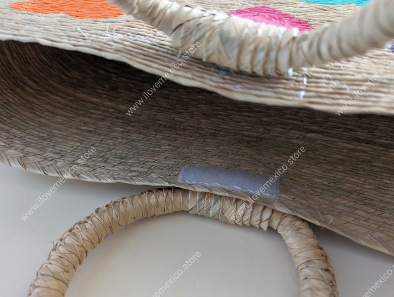May include: A woven straw tote bag featuring two round, rigid handles and a small rectangular velcro closure strip on the interior rim. The exterior surface displays embroidered floral patterns in bright orange, pink, and turquoise thread.