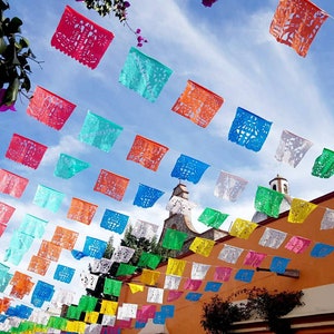 May include: Colorful paper flags strung across a street, creating a festive atmosphere. The flags are in various shapes and sizes, with intricate cutouts and designs. The flags are hung against a bright blue sky.