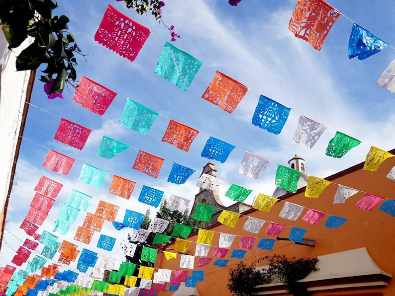May include: Colorful paper flags strung across a street, creating a festive atmosphere. The flags are in various shapes and sizes, with intricate cutouts and designs. The flags are hung against a bright blue sky.