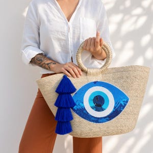 May include: A woven straw tote bag featuring a large, sequined evil eye motif in shades of blue, white, and black. The bag has two round, rigid straw handles and a decorative, multi-layered royal blue tassel hanging from one side of the handle.