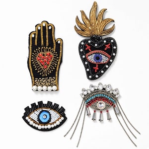 May include: Four decorative patches featuring intricate beadwork and embroidery. One patch is a hand with a red heart, one is a heart with a flame and eye, one is a stylised eye, and one is an eye with dangling chains and clear crystal accents.