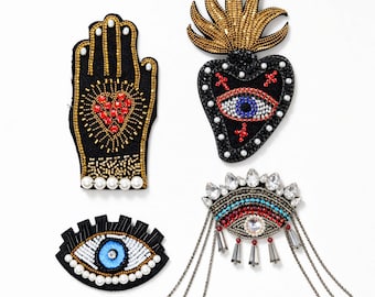 Op de afbeelding: Vier decoratieve patches met ingewikkeld kralenwerk en borduurwerk. Eén patch is een hand met een rood hart, één is een hart met een vlam en oog, één is een gestileerd oog en één is een oog met bungelende kettingen en heldere kristallen.