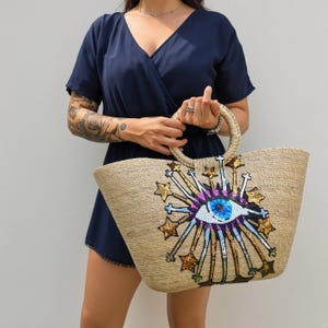 May include: A woven straw tote bag featuring a large, central eye motif embellished with sequins in shades of blue, purple, and black. The design includes radiating lines and gold sequined stars. The bag has two round, braided straw handles.