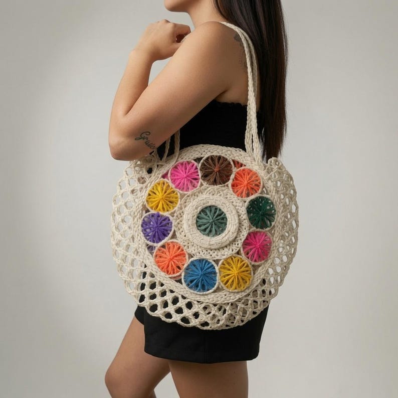 Pu&ograve; includere: Una borsa tote rotonda all'uncinetto color crema con un motivo circolare centrale circondato da piccoli accenti di filato colorati a forma di stella nelle tonalit&agrave; rosa, arancione, giallo, viola, blu, verde e marrone, con un design a rete aperta.
