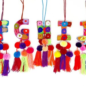 May include: Five colourful felt letter ornaments with pom-pom tassels. The letters are F, G, H, I, and J. Each letter is decorated with colourful embroidery and has a string for hanging.