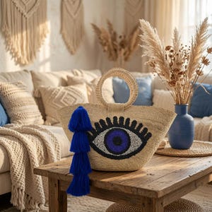 Op de afbeelding: Een geweven strooien tas met een rond handvat en een opvallend geborduurd boze oog-motief in zwart, wit en blauw. Een lange, gelaagde koningsblauwe kwast hangt aan de zijkant van de tas en voegt een levendig accent toe aan het ontwerp.