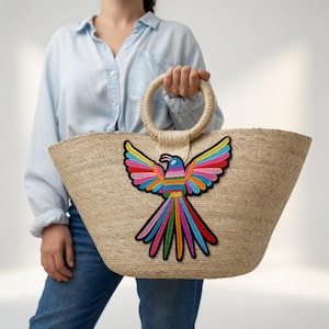 May include: A woven straw tote bag featuring a large, colorful embroidered parrot with outstretched wings. The bag has a circular straw handle and a wide, tapered body. The parrot design incorporates vibrant shades of red, blue, yellow, and green.