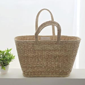 May include: A woven straw tote bag with two handles. The bag is rectangular with a wide opening and a textured surface. The handles are also woven and attached to the top of the bag. A small potted plant is to the left of the bag.