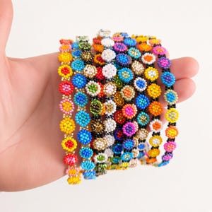 Puede incluir: Una colección de varias pulseras de cuentas con pequeños patrones florales redondos y multicolores. Cada pulsera está hecha de diminutas cuentas de vidrio en tonos vibrantes de amarillo, azul, rosa, verde, naranja, blanco y negro.