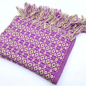 May include: A folded rectangular textile featuring a repeating geometric diamond pattern in vibrant purple and lime green. The fabric is finished with a decorative fringe along one edge, composed of twisted strands of both purple and lime green yarn.
