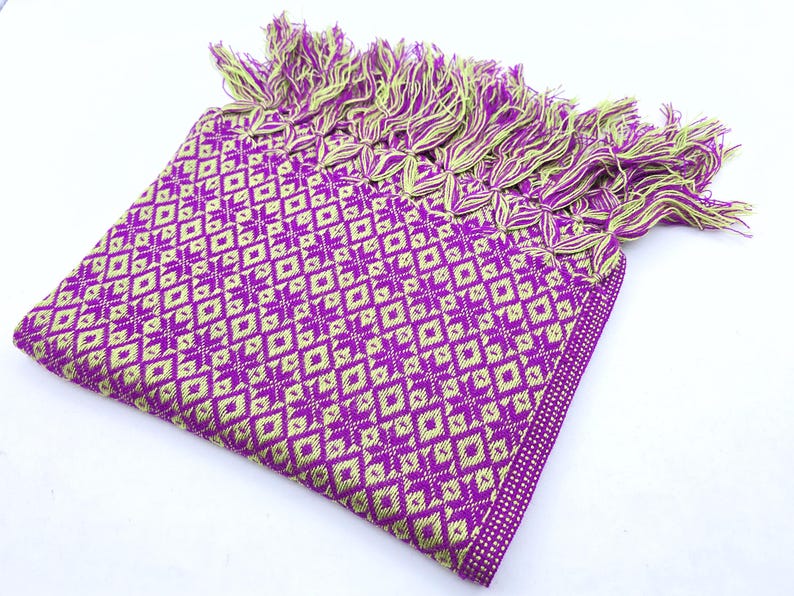 May include: A folded rectangular textile featuring a repeating geometric diamond pattern in vibrant purple and lime green. The fabric is finished with a decorative fringe along one edge, composed of twisted strands of both purple and lime green yarn.