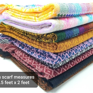 May include: A stack of folded woven scarves featuring various geometric patterns and colors including yellow, pink, blue, gold, black, and maroon. Each scarf measures 6.5 feet by 2 feet and features fringed ends on one side of the fabric stack.