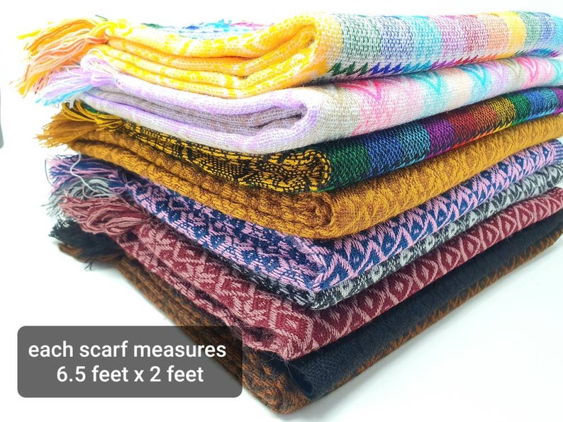 May include: A stack of folded woven scarves featuring various geometric patterns and colors including yellow, pink, blue, gold, black, and maroon. Each scarf measures 6.5 feet by 2 feet and features fringed ends on one side of the fabric stack.