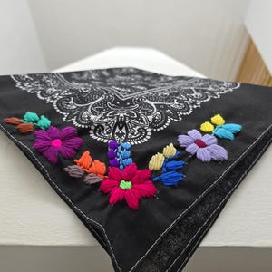 May include: A black bandana featuring a white paisley print pattern and colorful floral embroidery. The embroidery includes bright pink, purple, yellow, blue, and orange thread, creating a vibrant contrast against the dark fabric background.