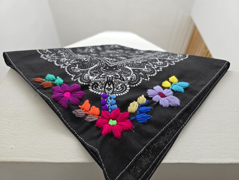 May include: A black bandana featuring a white paisley print pattern and colorful floral embroidery. The embroidery includes bright pink, purple, yellow, blue, and orange thread, creating a vibrant contrast against the dark fabric background.