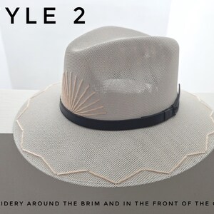 May include: A light beige straw fedora hat featuring a black leather band, a sunburst embroidery design on the front of the crown, and a zigzag embroidered pattern around the edge of the brim. The text STYLE 2 appears at the top of the image.
