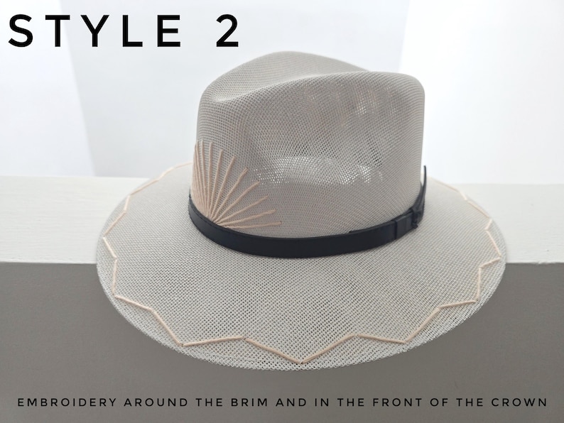 May include: A light beige straw fedora hat featuring a black leather band, a sunburst embroidery design on the front of the crown, and a zigzag embroidered pattern around the edge of the brim. The text STYLE 2 appears at the top of the image.