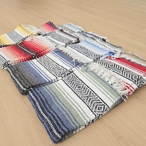 May include: A collection of folded woven blankets featuring various color gradients including red, yellow, blue, and green. Each blanket displays a central black and white geometric diamond pattern, striped borders, and frayed fringe edges on the ends.