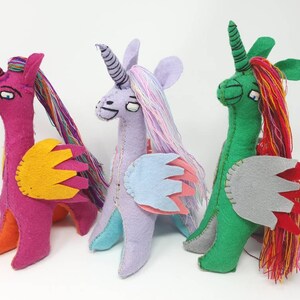 Puede incluir: Tres peluches de unicornio hechos a mano en fieltro dispuestos en fila. El primero es magenta con alas amarillas y pelo de hilo multicolor. El segundo es lavanda con alas azul claro. El tercero es verde con alas grises y pelo de hilo rojo.
