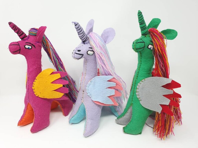 Puede incluir: Tres peluches de unicornio hechos a mano en fieltro dispuestos en fila. El primero es magenta con alas amarillas y pelo de hilo multicolor. El segundo es lavanda con alas azul claro. El tercero es verde con alas grises y pelo de hilo rojo.