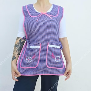 May include: A purple and pink gingham apron with white lace trim and pink embroidered flowers. The apron has two pockets and ties at the waist.