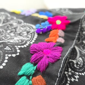May include: A black fabric item featuring a white paisley mandala pattern and vibrant, multi-colored floral embroidery. The embroidery includes bright pink, orange, purple, blue, and green thread, creating textured, raised flower designs.