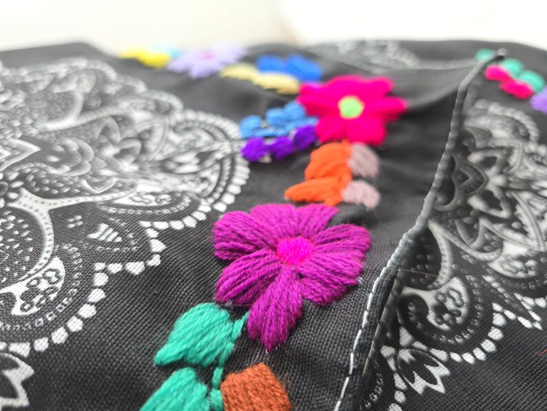 May include: A black fabric item featuring a white paisley mandala pattern and vibrant, multi-colored floral embroidery. The embroidery includes bright pink, orange, purple, blue, and green thread, creating textured, raised flower designs.