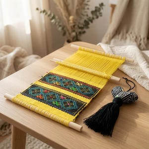 May include: A yellow woven wall hanging featuring two horizontal bands of intricate geometric embroidery in black, blue, green, and orange. The textile is suspended between two light-colored wooden dowels and includes a black heart-shaped tassel.