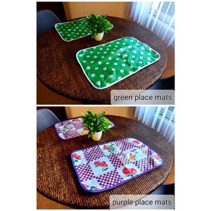 May include: Two placemats on a table. One placemat is green with white polka dots and the other is white with a purple border and a red and green fruit pattern. The text "green place mats" and "purple place mats" is below each placemat.