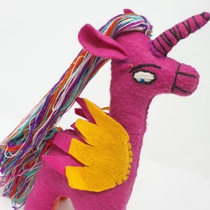 Puede incluir: Un peluche de unicornio hecho a mano en fieltro de color magenta vibrante. Presenta una larga melena de hilo multicolor, alas de fieltro amarillo y un cuerno en espiral con costuras negras. El rostro tiene ojos y hocico bordados a mano.