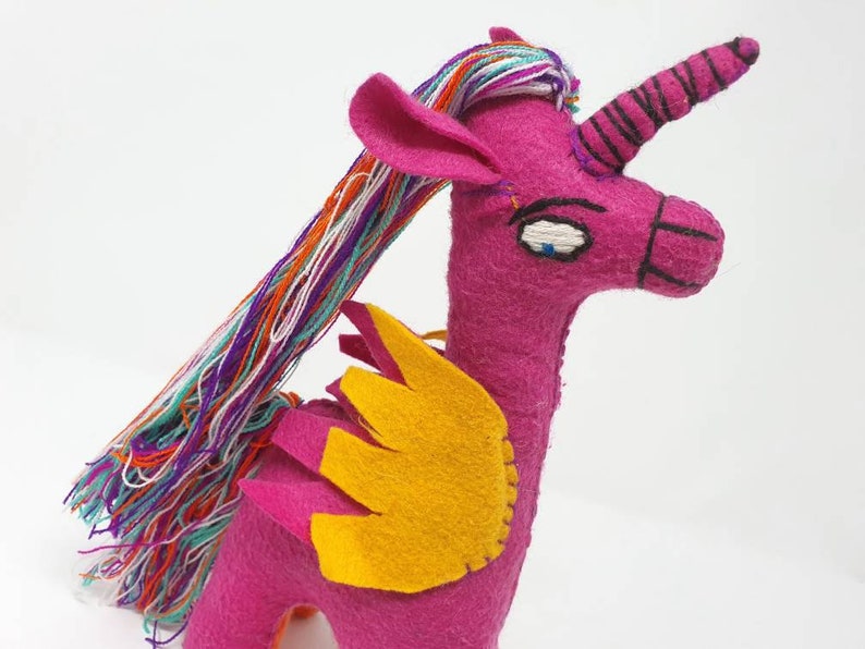 Puede incluir: Un peluche de unicornio hecho a mano en fieltro de color magenta vibrante. Presenta una larga melena de hilo multicolor, alas de fieltro amarillo y un cuerno en espiral con costuras negras. El rostro tiene ojos y hocico bordados a mano.
