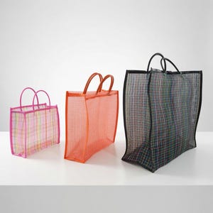 May include: Three rectangular tote bags made of a semi-transparent mesh material. The smallest bag is pink with a plaid pattern, the middle bag is solid orange, and the largest bag is black with a multi-colored grid pattern and black trim.