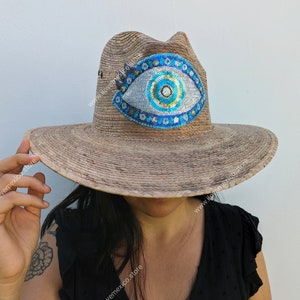 Puede incluir: Un sombrero de paja de ala ancha con un diseño de ojo turco azul y dorado. El sombrero está hecho de paja natural y tiene un aspecto desgastado.