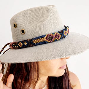 Peut inclure: Un chapeau fedora beige clair à larges bords doté de deux œillets métalliques couleur laiton sur le côté. Le chapeau est orné d'une bande tissée à motifs géométriques bleu foncé, bordeaux et dorés, avec des cordons tressés pendants.