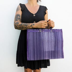 May include: A rectangular tote bag made of a semi-transparent purple mesh material featuring vertical stripes in varying shades of purple and white. The bag has a reinforced top border with silver-toned metal rivets and two sturdy dark handles.